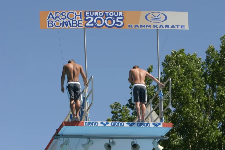 Zwei Springer auf der Plattform unter dem Schild Arschbombe Eurotour 2005 vor blauem Himmel.