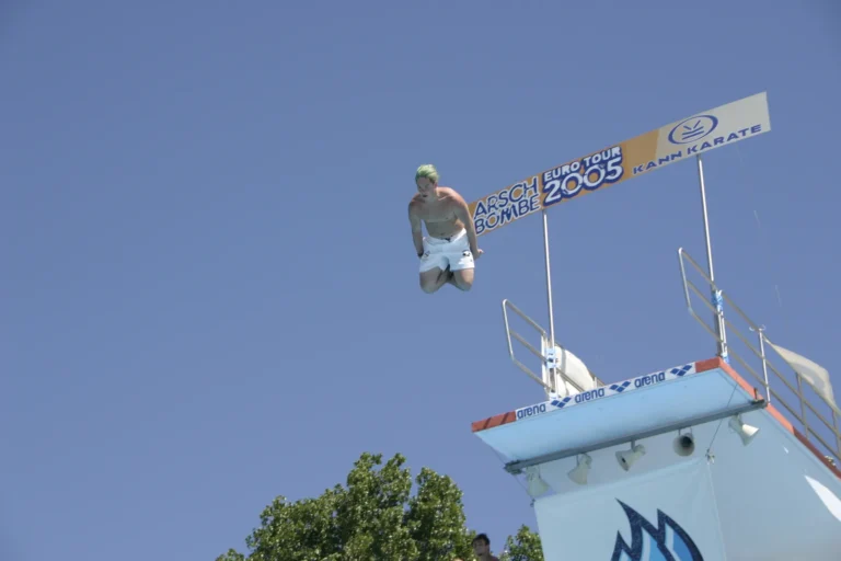 Ein Springer mit angewinkelten Beinen in der Luft neben dem Arschbombe-Eurotour-2005-Schild.
