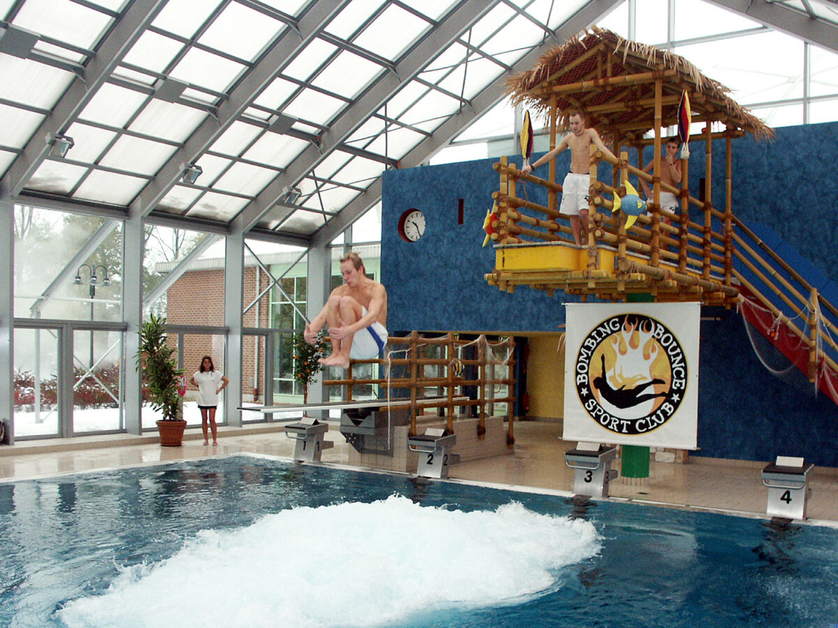 Ein Springer in kompakter Hocke über dem Becken, dahinter der Sprungturm und das Bombing-Bounce-Sport-Club-Banner im Bahia in Bocholt, 2005.