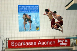 Two divers in red shorts airborne together inside an indoor pool, 2005.