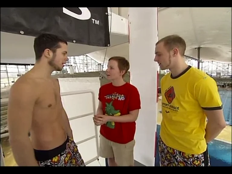 Ein junger Reporter im roten Shirt spricht am Hallenbecken mit zwei Springern.